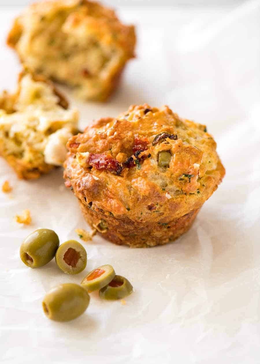 Muffins de queso salado con tomates con sol, queso feta y aceitunas en papel blanco, con aceitunas a un lado, listas para ser comidas.